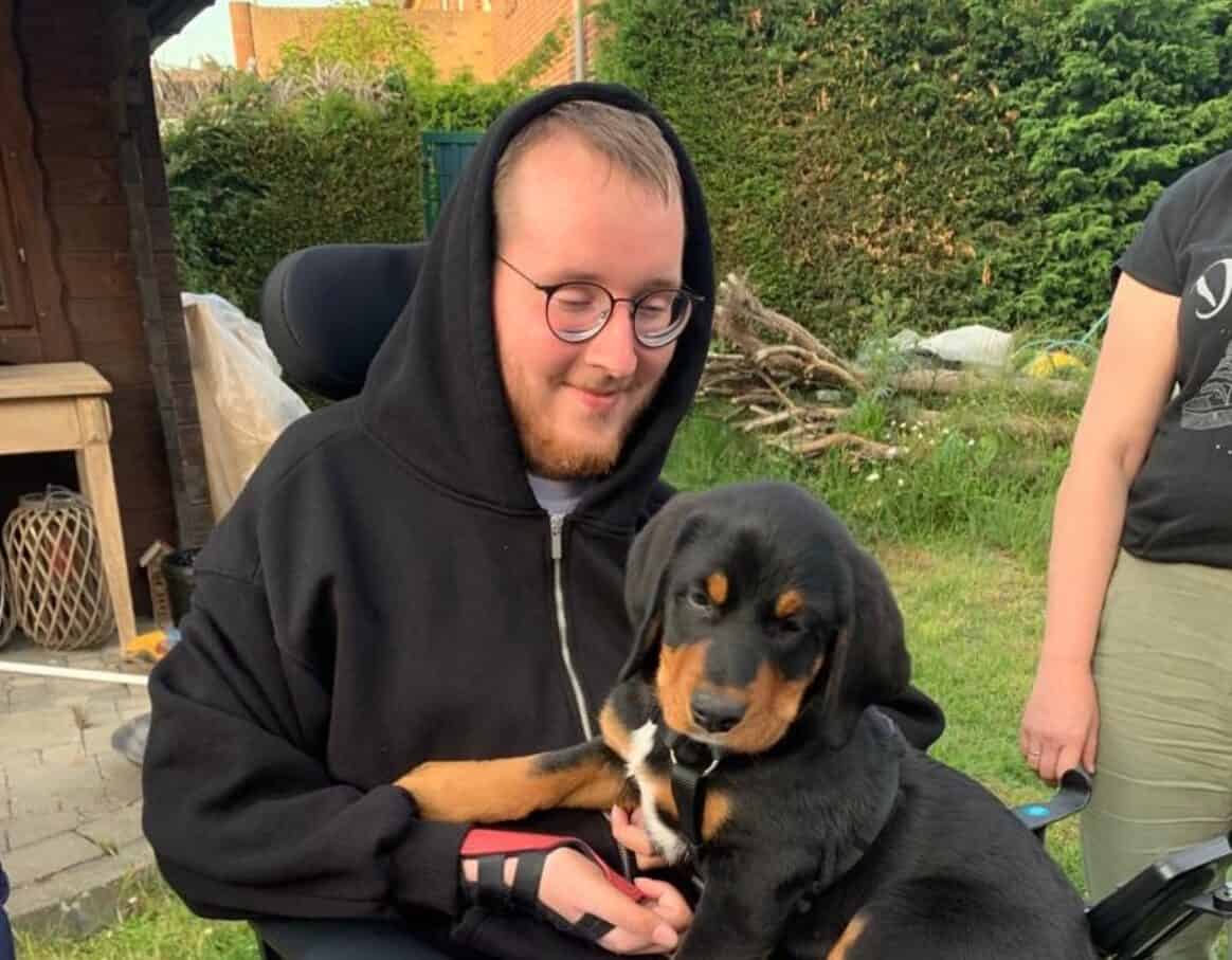 Lukas im schwarzen Hoodie im E-Rollstuhl im Garten mit einem seiner Hunde auf dem Schoß.