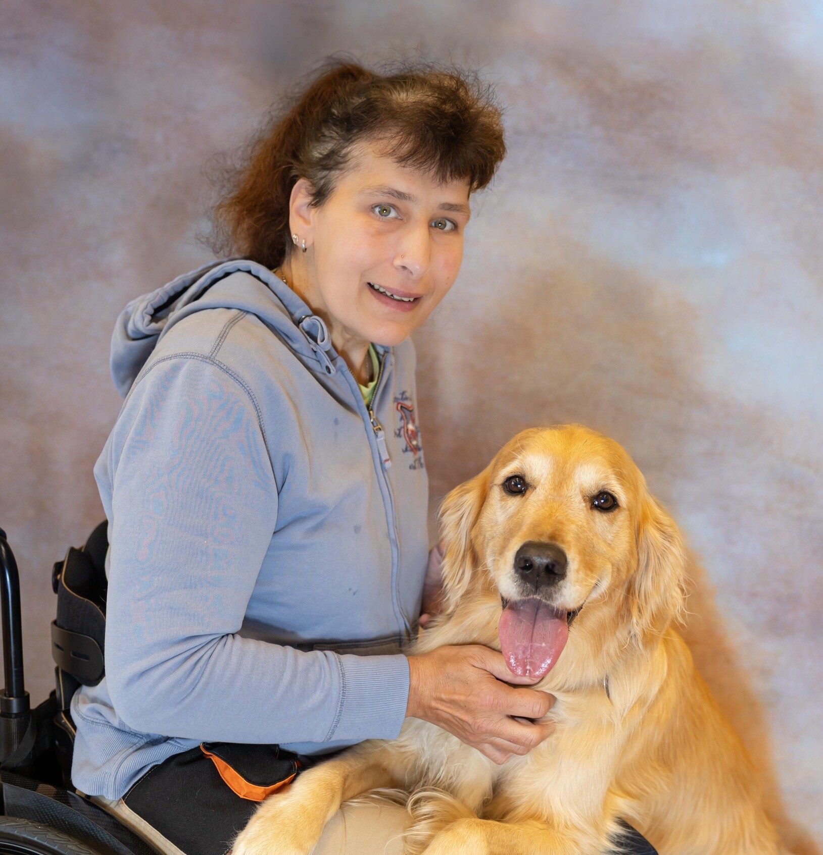 Eine weiblich gelesene Person mit Kapuzenpulli im Rollstuhl. Ein Retriever Hund hat seine Vorderpfoten auf ihrem Schoß abgestellt.