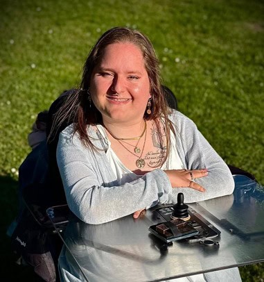 Eine lächelnde Person sitzt draußen auf einer Wiese an einem Rollstuhltisch. Sie hat lange blonde Haare. Im Hintergrund grünes Gras.