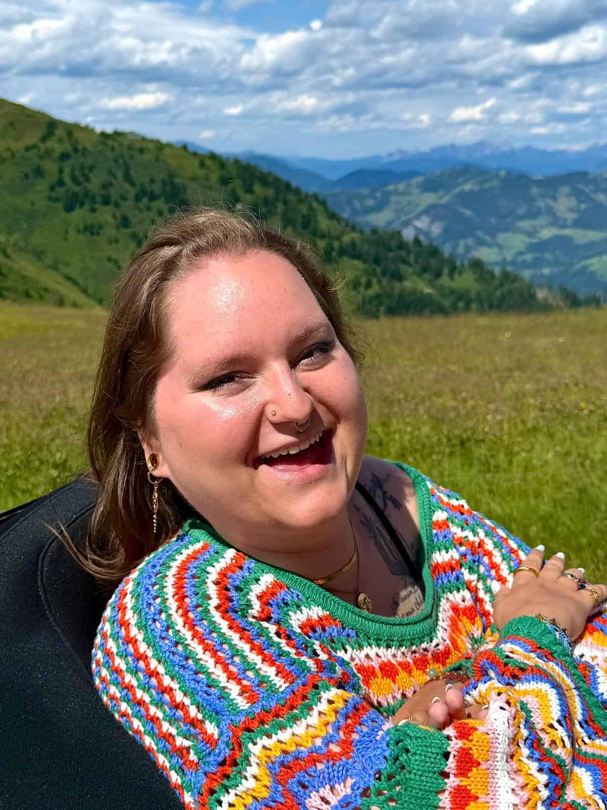 Eine weiblich gelesene Person im bunt gestrickten Pullover lächelt bei Sonnenschein in die Kamera.