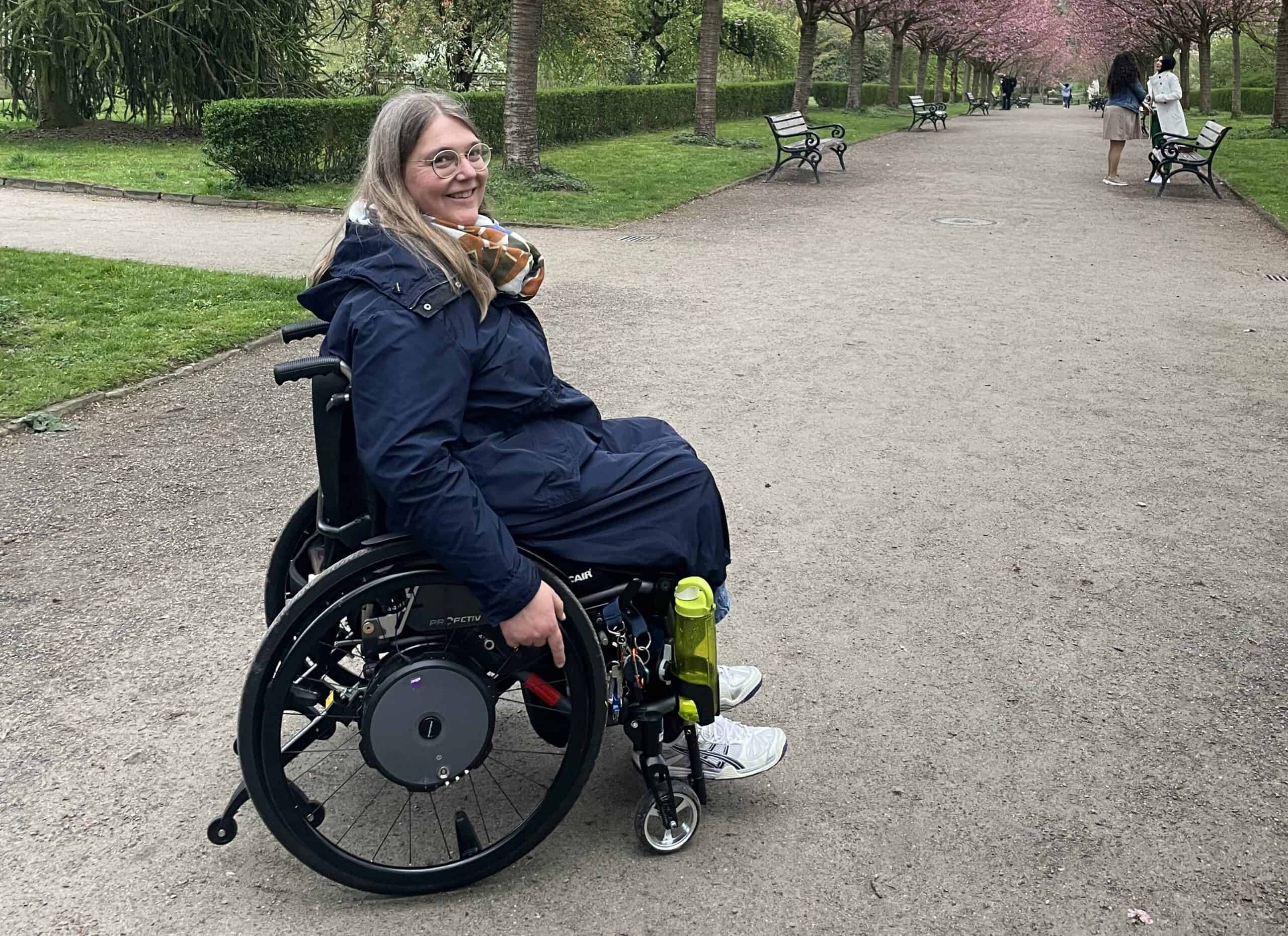 Anja lächelt in die Kamera. Sie sitzt mit den Händen am Rollstuhl, bereit, den Parkweg runterzufahren.
