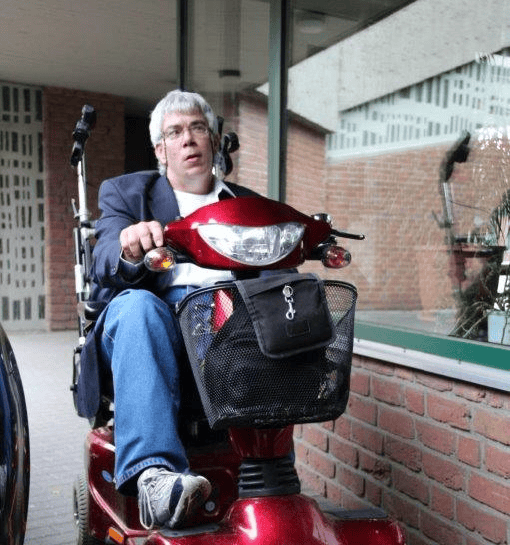 Klaus fährt auf seinem roten Roller. Er trägt sportliche Schuhe und eine Brille.