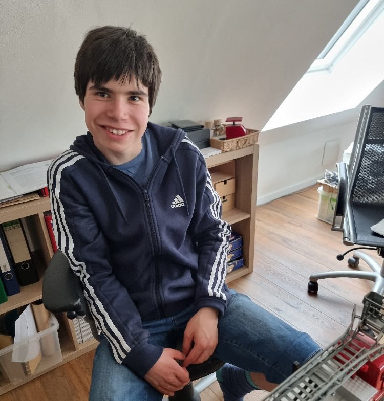 Ein 19-jähriger junger Mann sitzt locker auf einem Stuhl am Schreibtisch und lächelt.