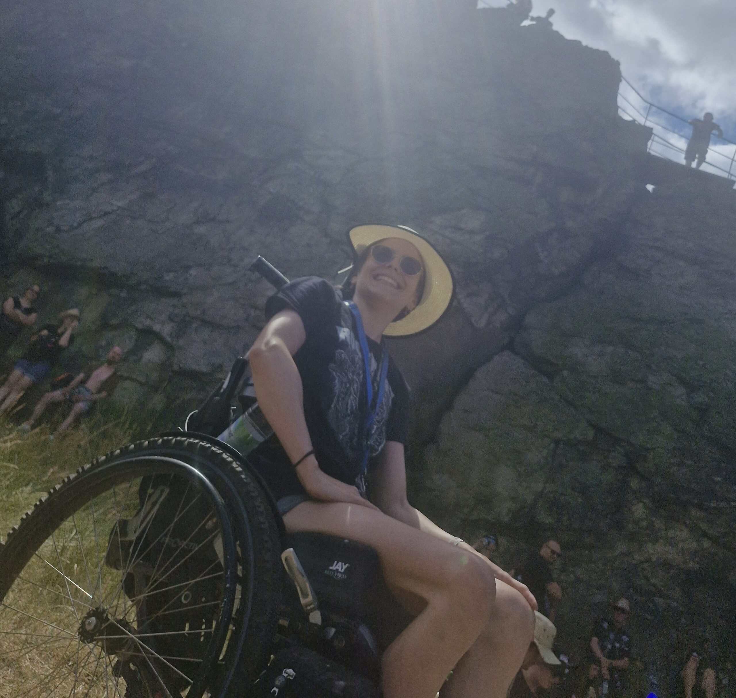 Julia sitzt grinsend in ihrem Rollstuhl vor einer Felsenwand. Die Sonne scheint ihr ins Gesicht und sie hat eine Sonnenbrille und einen Strohhut auf.