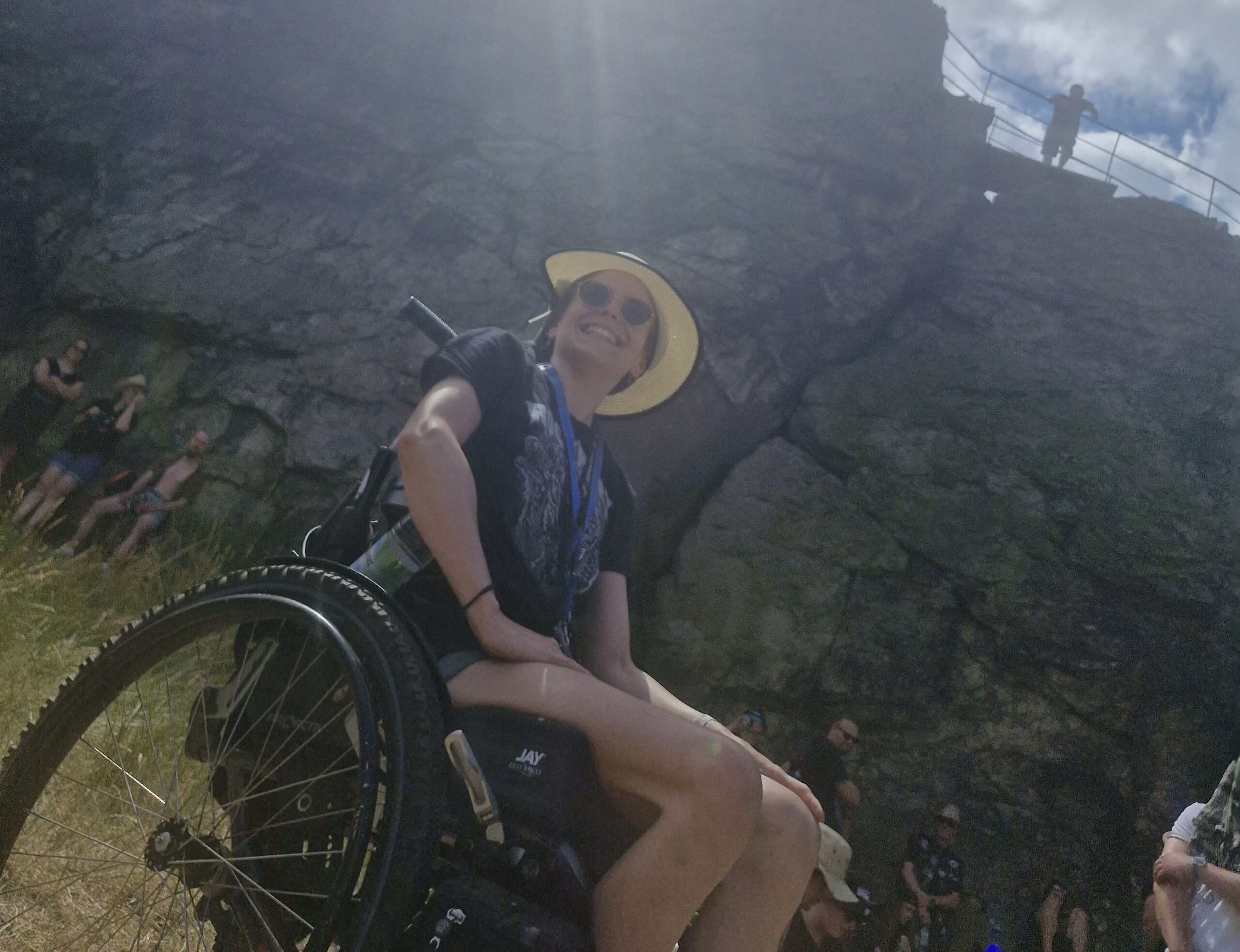 Julia sitzt grinsend in ihrem Rollstuhl vor einer Felsenwand. Die Sonne scheint ihr ins Gesicht und sie hat eine Sonnenbrille und einen Strohhut auf.