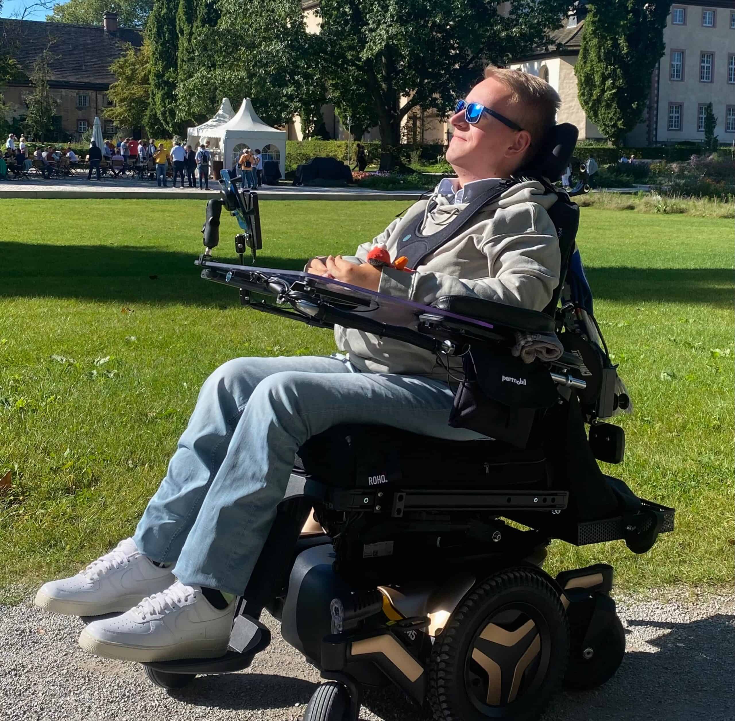 Ein Mann mit Sonnenbrille sitzt im Rollstuhl im Grünen.