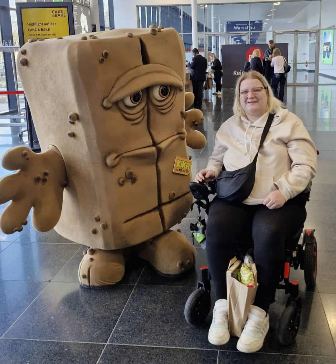 Lena sitzt in ihrem elektrischen Rollstuhl neben der bekannten KiKA-Figur „Bernd das Brot“. Beide befinden sich in einer hellen Messehalle. Lena trägt einen hellen Hoodie, schwarze Hose und weiße Sneaker und lächelt in die Kamera. Auf der Fußablage ihres Rollstuhls steht eine Einkaufstüte. Im Hintergrund sind weitere Menschen sowie verschiedene Messestände zu sehen