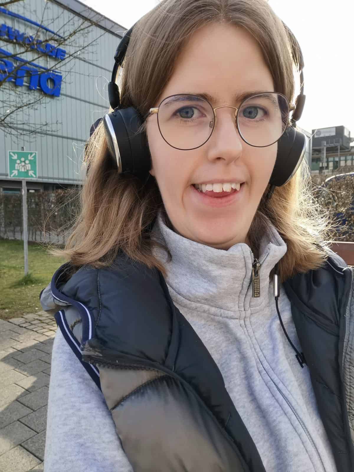 Eine Person mit schulterlangen Haaren, Brille und Kopfhörern lächelt in die Kamera.